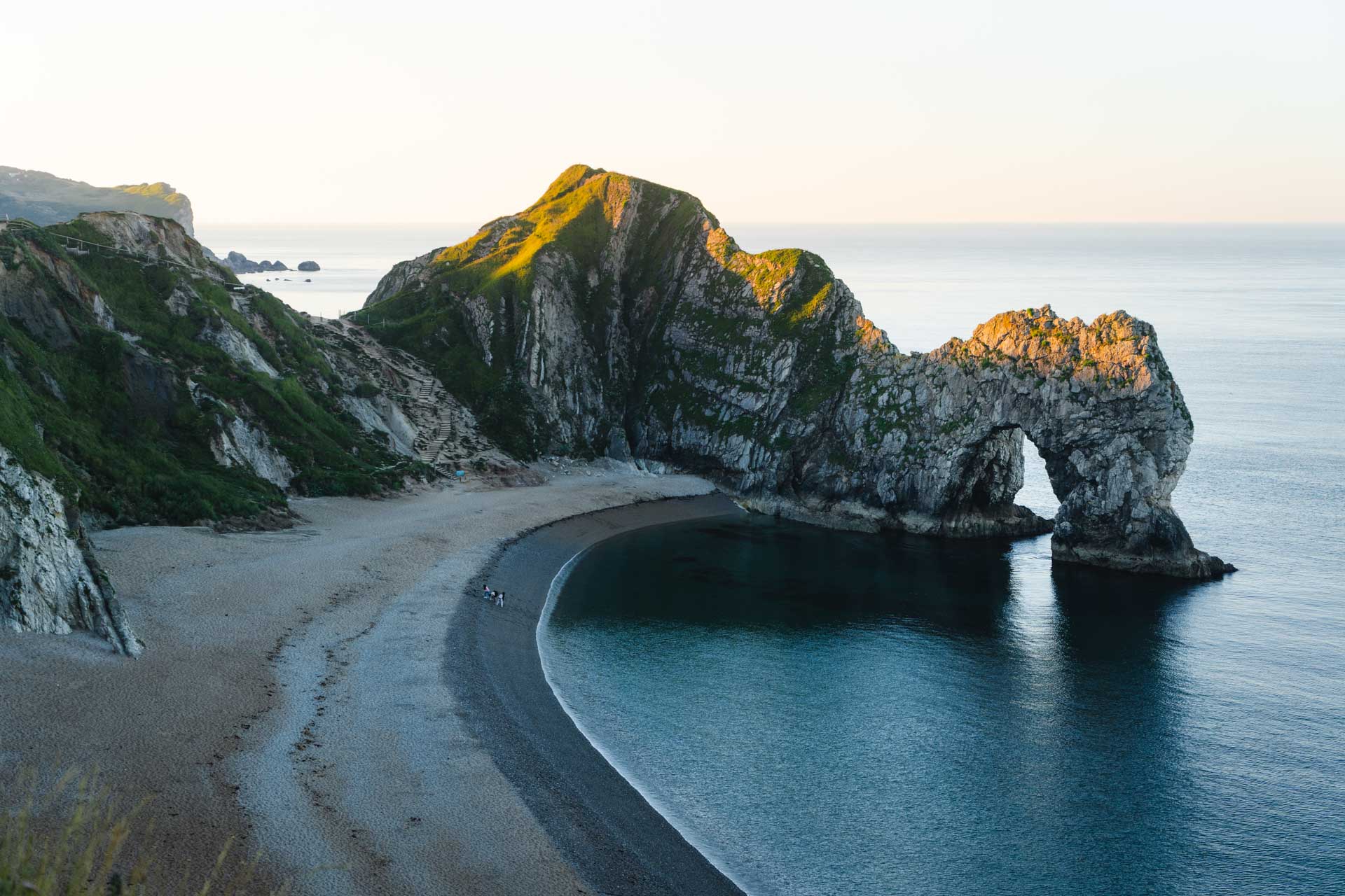 The Durdle Door rock formation in Dorset glowing in the sunrise light as two people look on from the beach.