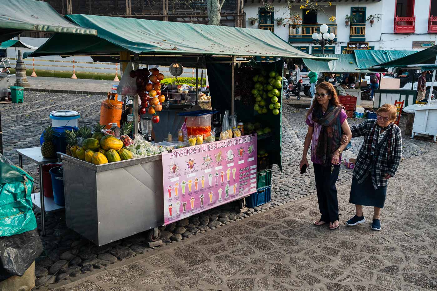 Two women walking past a fruit vendor in Jardin's Plaza de Libertador.