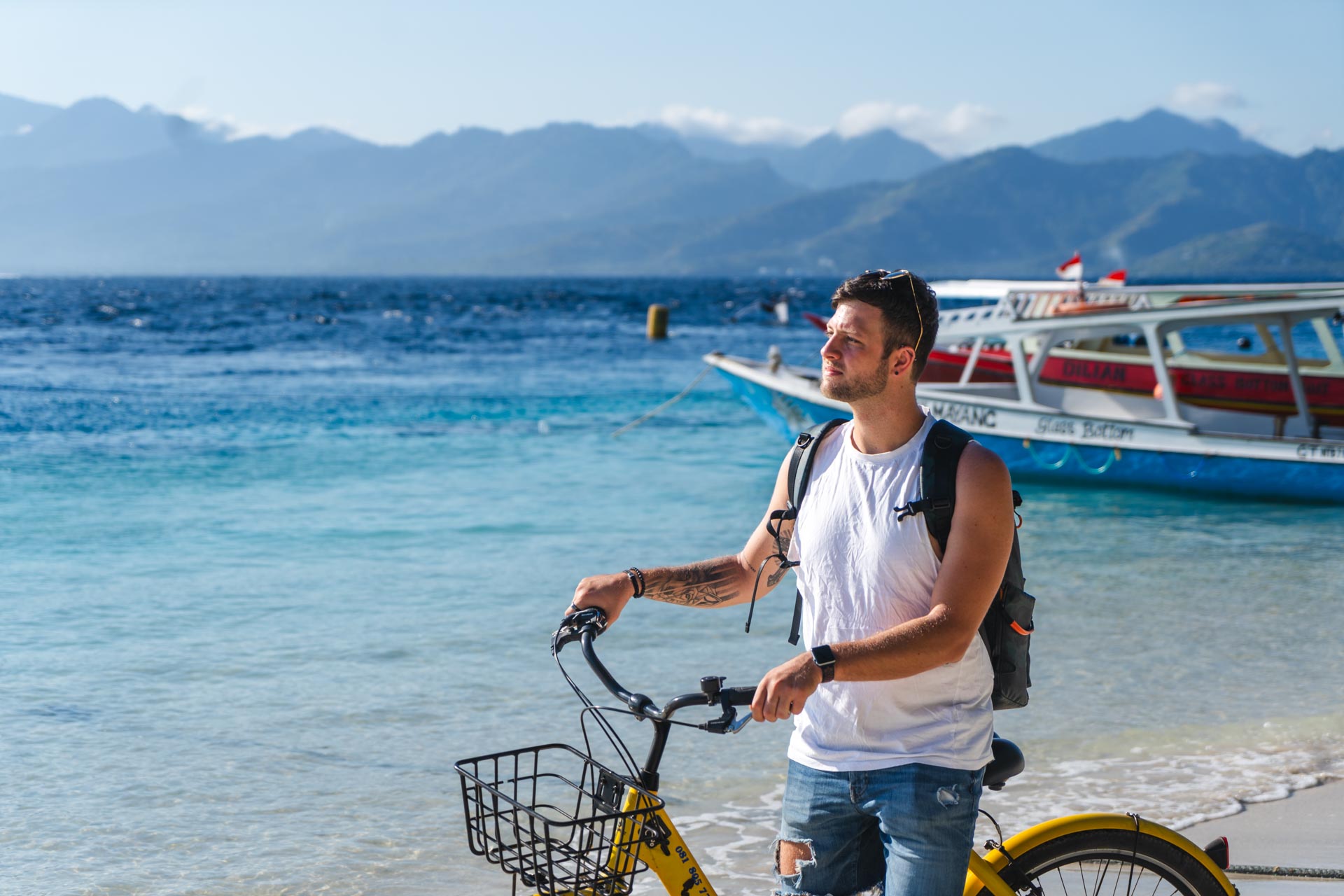 Ryan posing on a beach on Gili Trawangan island holding a bike besides crystal blue oceans.