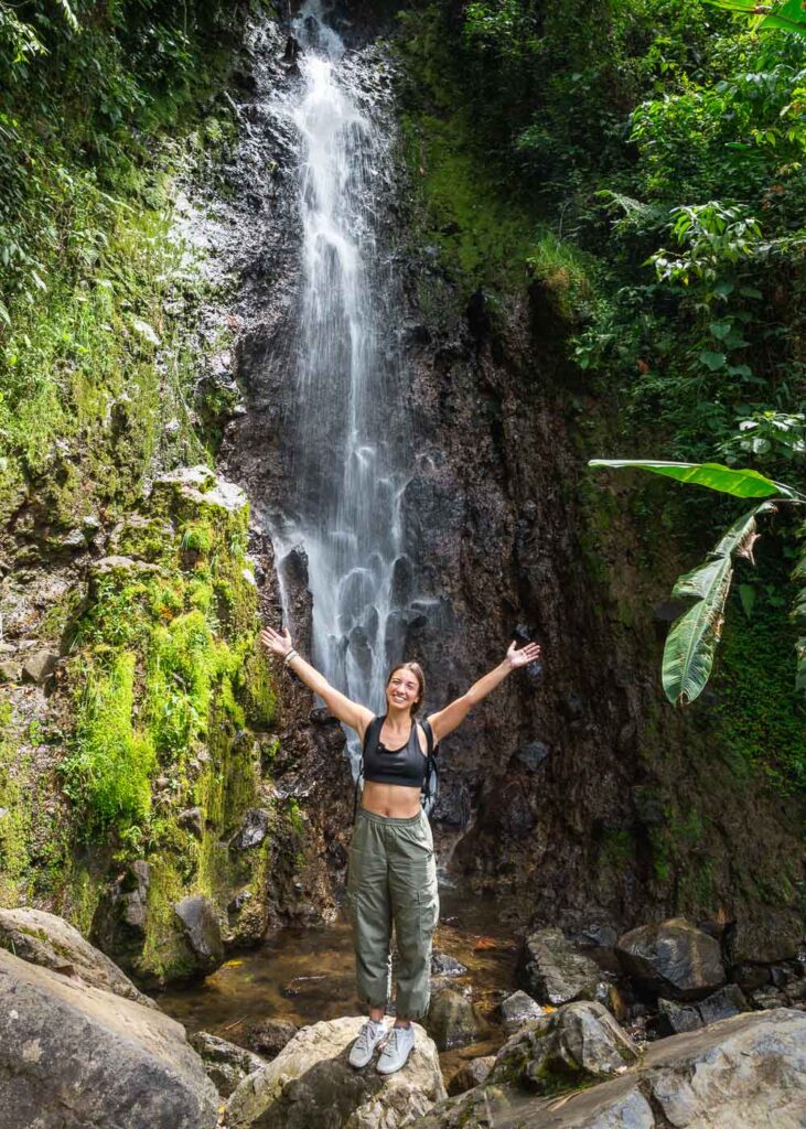 Sara smiling and posing on a rock with her hands in the air in front of Cascada del Amor.