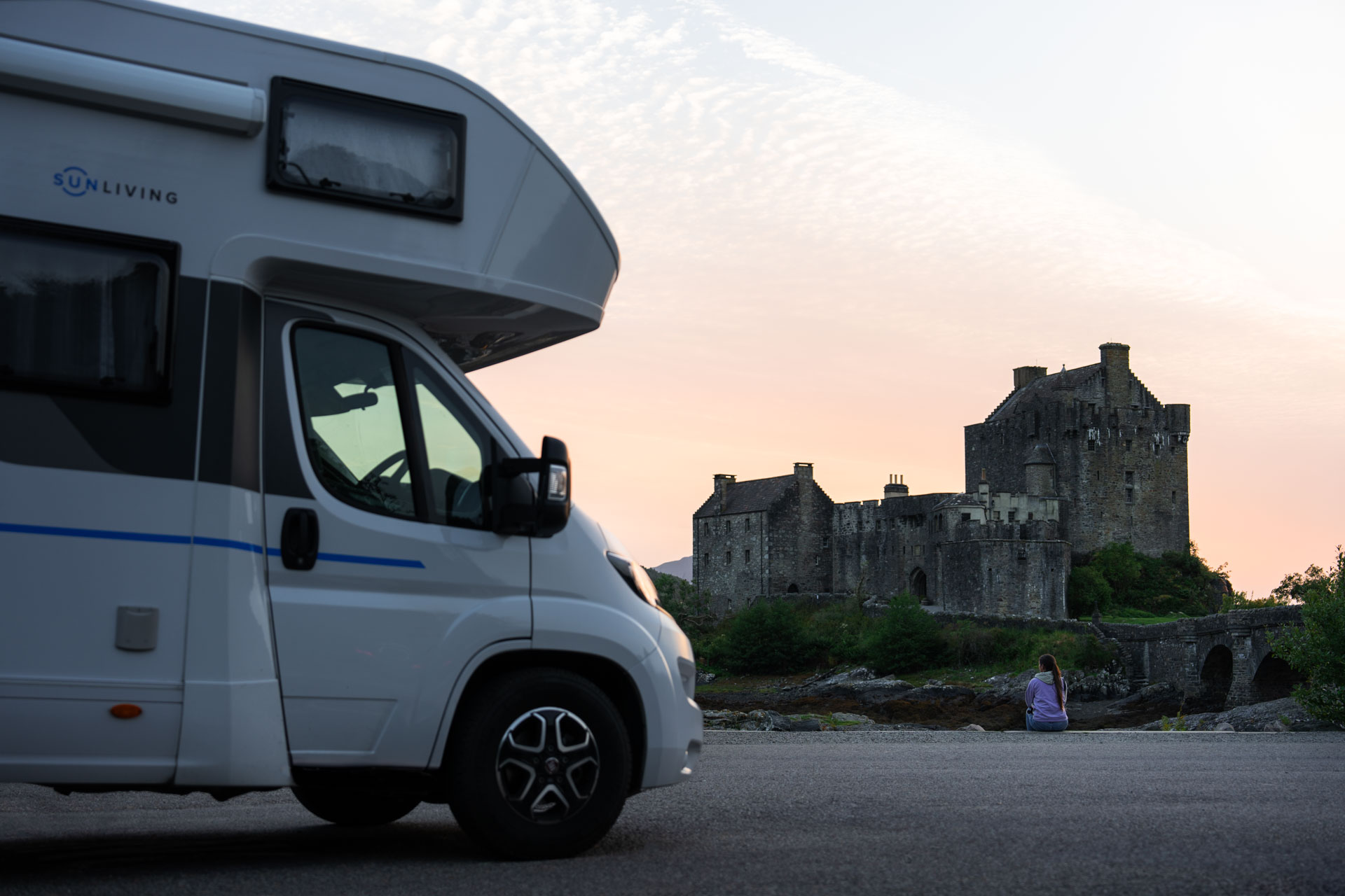 Sara sitting down on a wall enjoying the view of Eilean Donan Castle in Scotland besides a campervan.