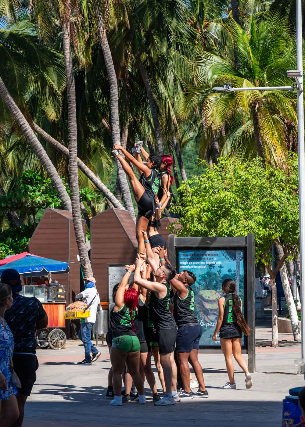 Cheerleaders wearing black and green clothes practicing on Rodadero promenade beside tourists and palm trees.
