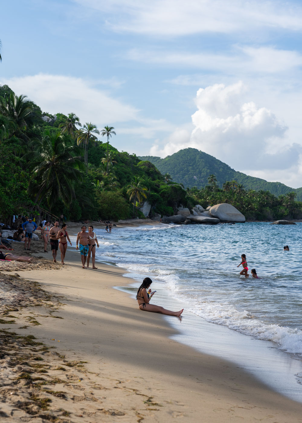 A girl on her phone sitting down on La Piscina beach in Tayrona National Park while other tourists go about their day.