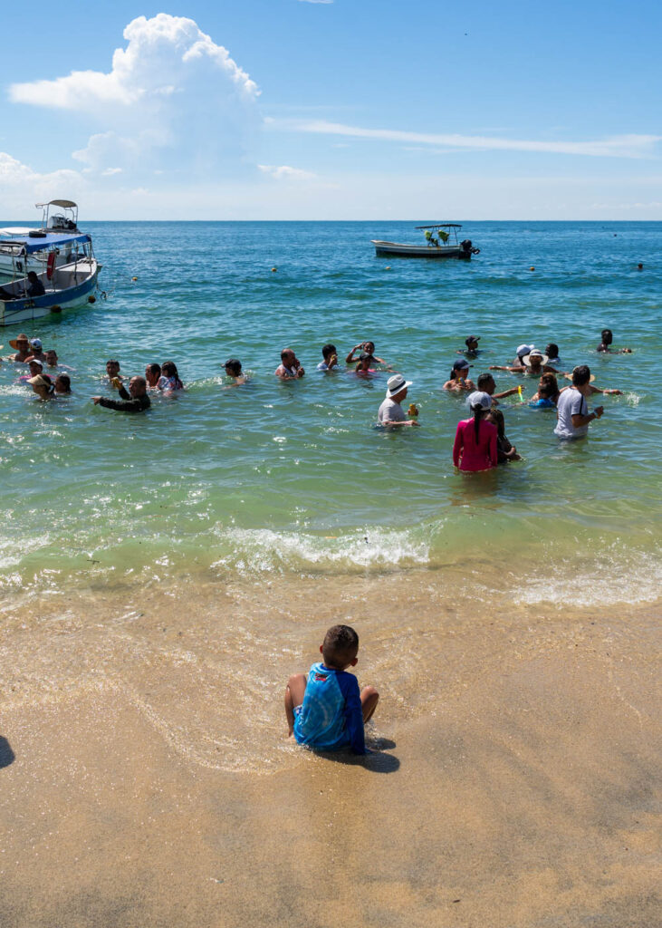 A young boy sitting on Playa Blanca watching people swim.