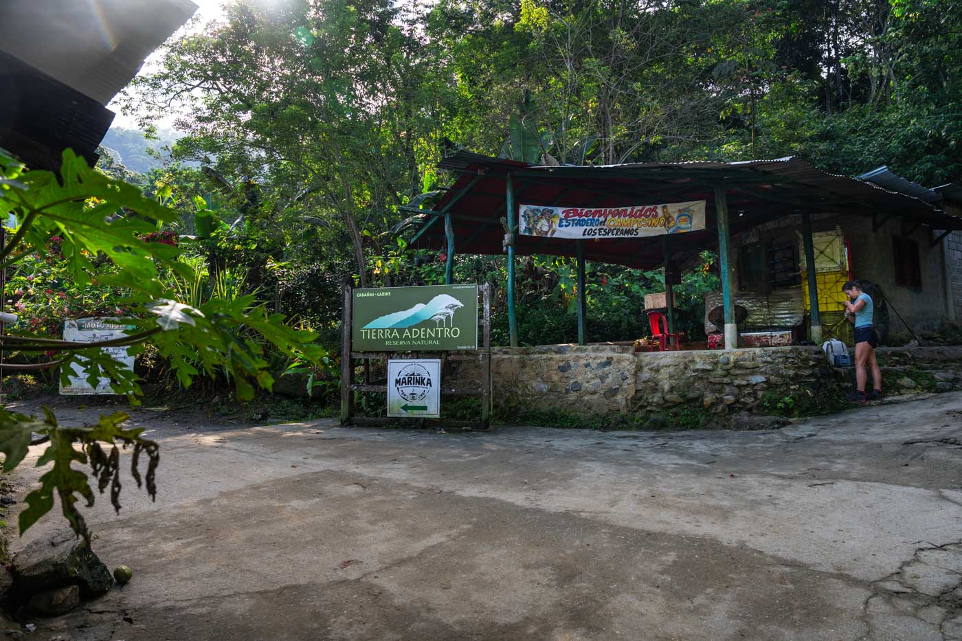 Sara standing besides a sheltered area at a fork in the road with a sign for Marinka Waterfalls on a gate.