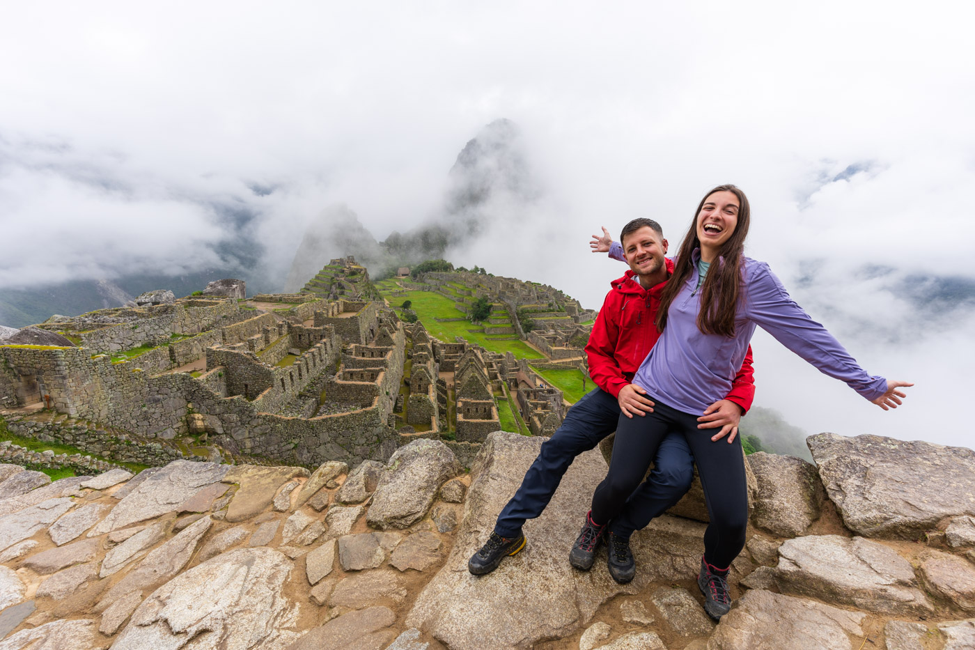 Ryan and Sara wearing colorful hiking gear and in a hugging pose sitting on a rock in front of a full view of Machu Picchu in Peru.
