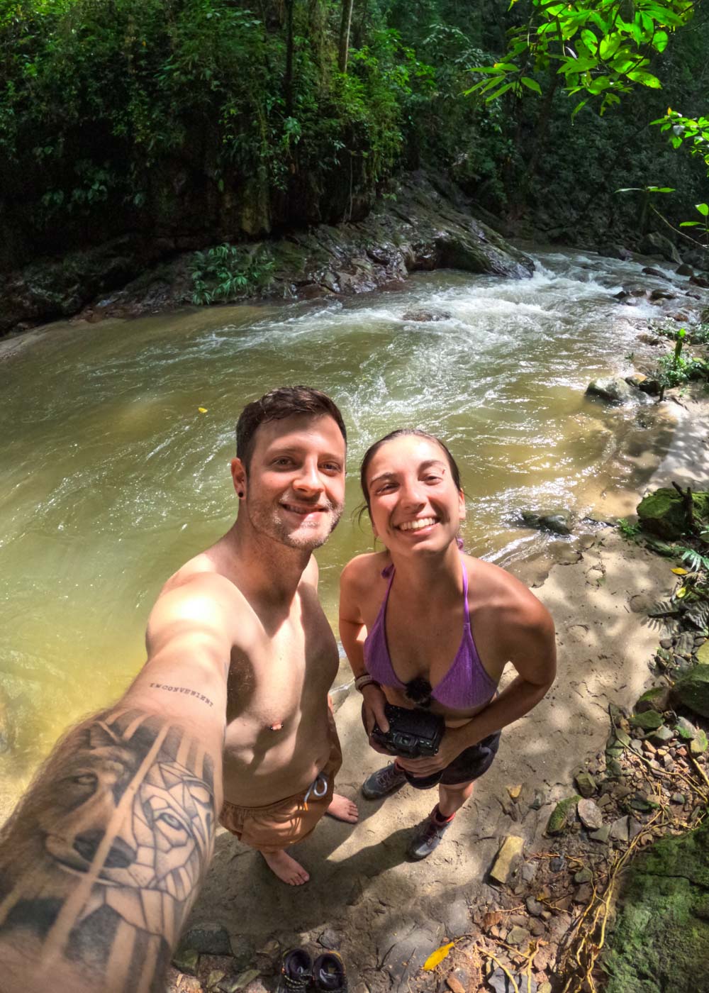 Ryan in an orange bathing suit and Sara in a purple bikini taking a selfie while standing besides a river in Minca.