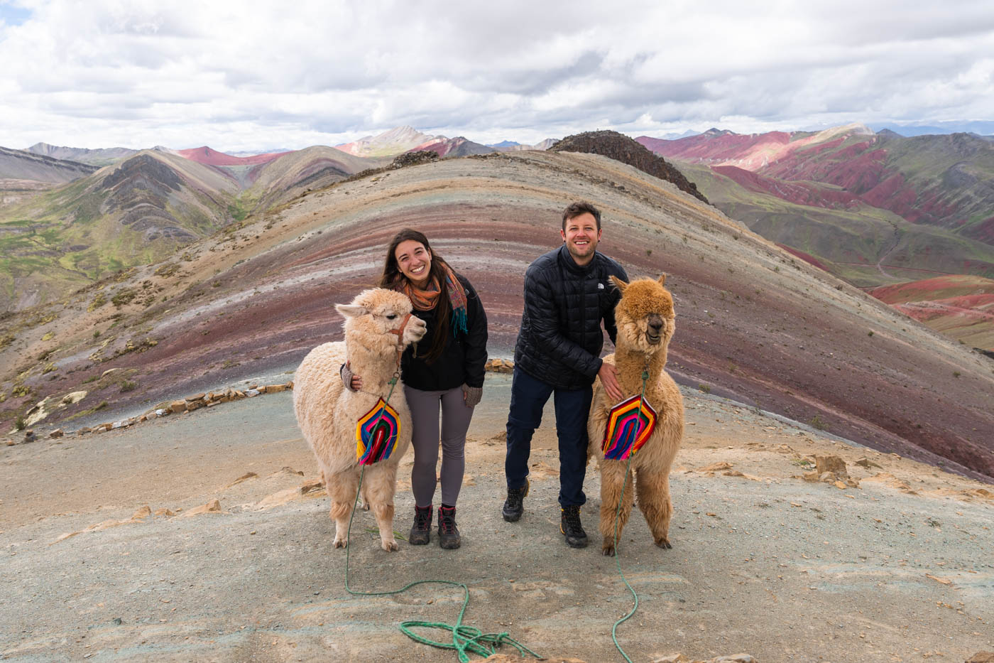 Ryan and Sara in hiking gear at the summit of Palcoyo rainbow mountain whilst hugging two fluffy alpacas.