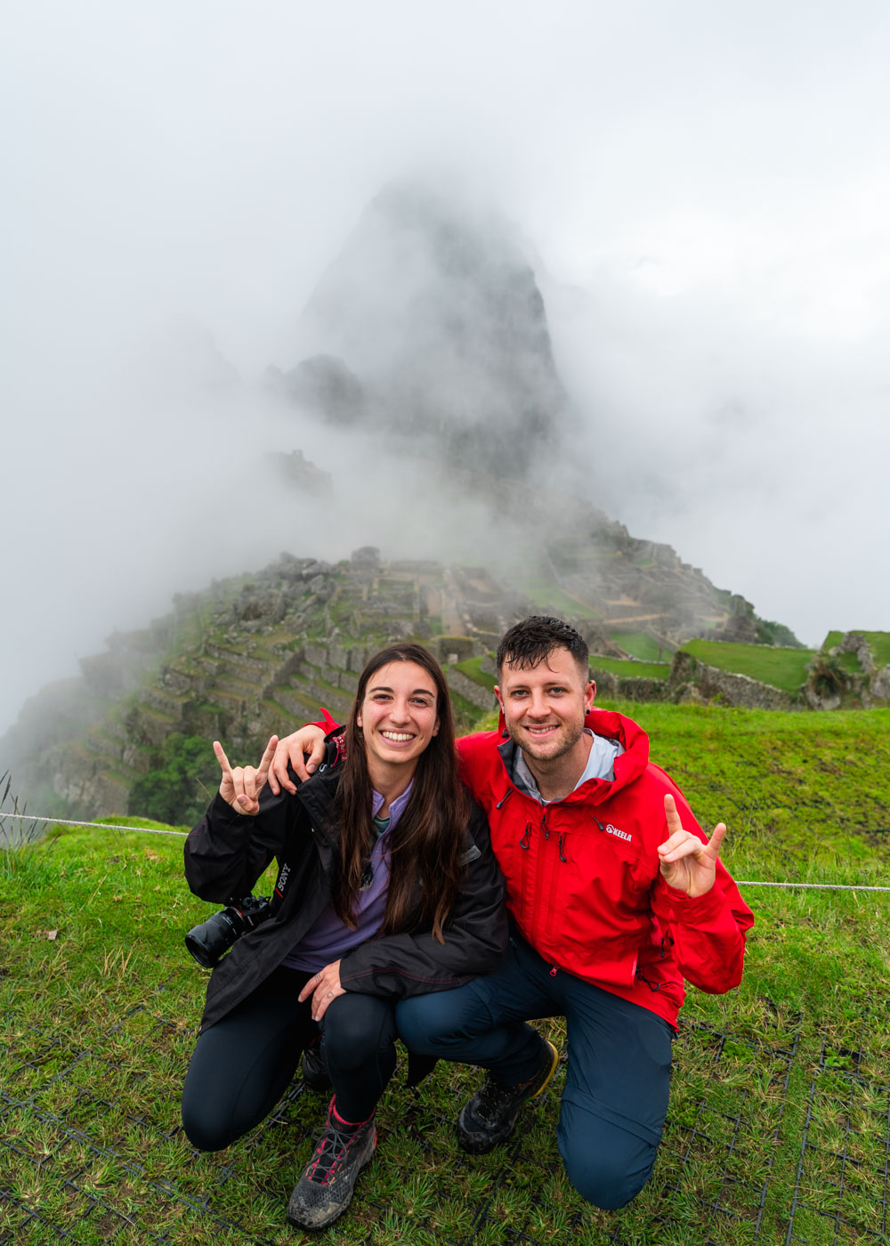 Ryan and Sara wet from the rain dressed in hiking gear posing in front of a cloudy Machu Picchu.