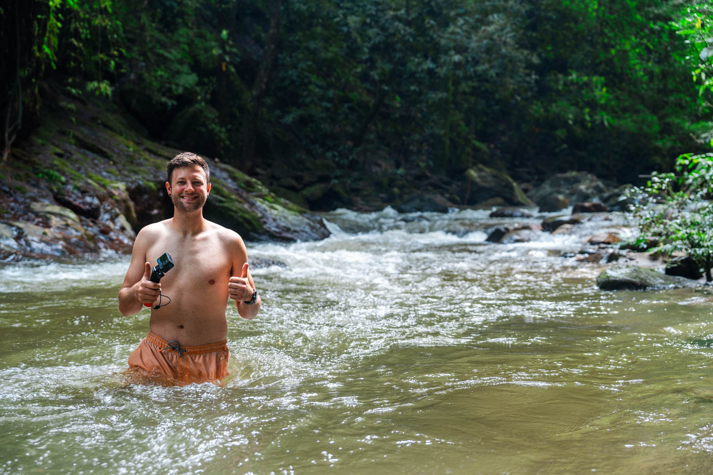 Ryan in orange swimmers and holding a GoPro standing waist deep in a river in Minca in the jungle.