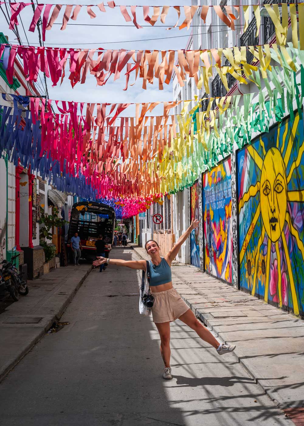 Sara making a silly pose in the middle of a street in Santa Marta historic center with rainbow colored decorations above.