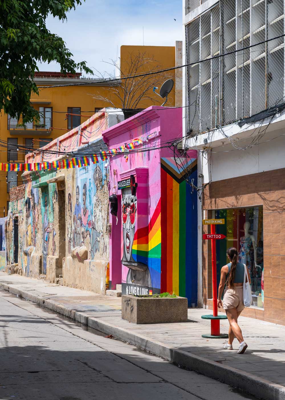 Sara walking down a colorful street past a oink and rainbow building on a sunny day in Santa Marta historic center.