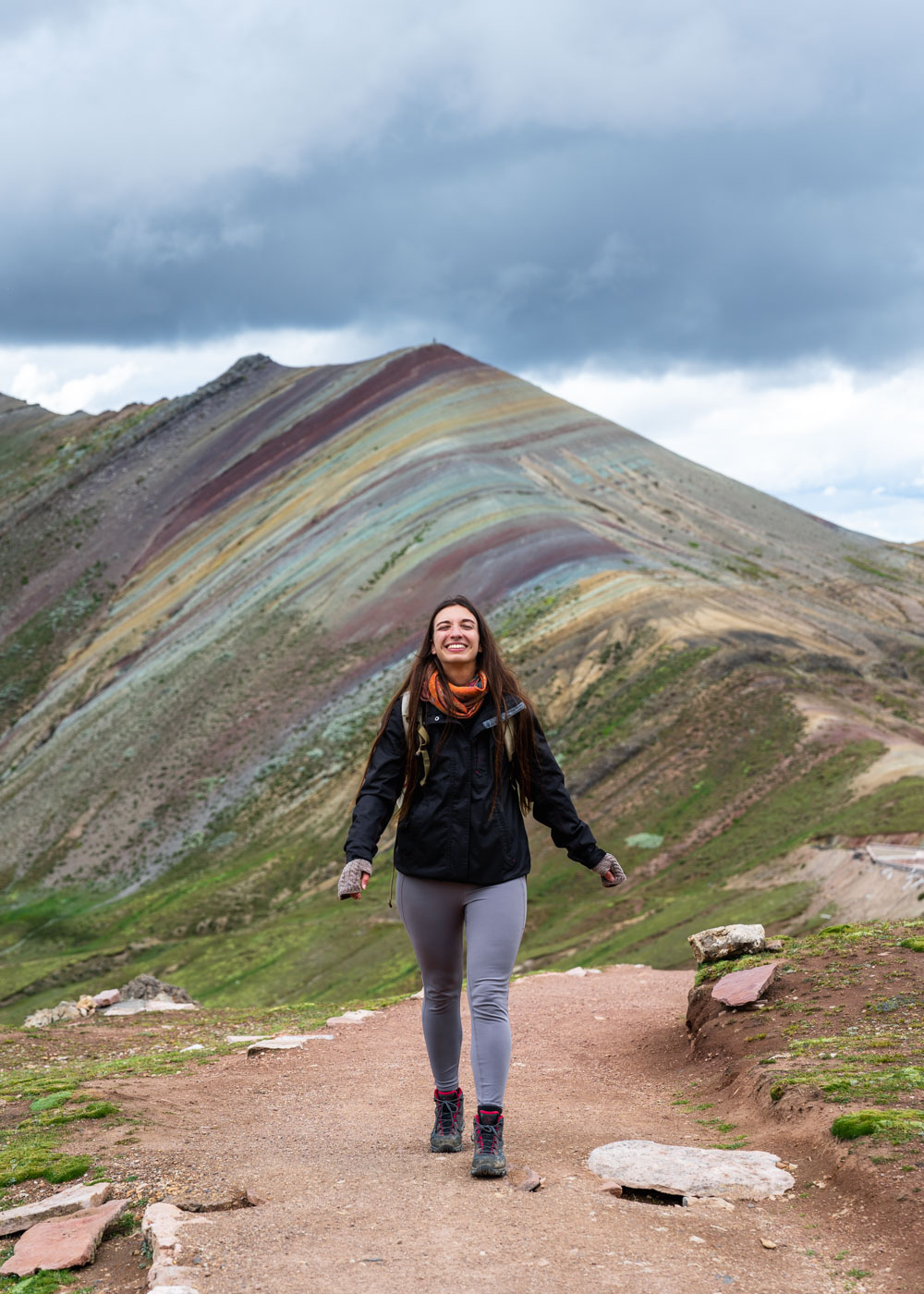 Sara in hiking gear smiling and posing in front of Palcoyo rainbow mountain in Peru with an overcast sky.