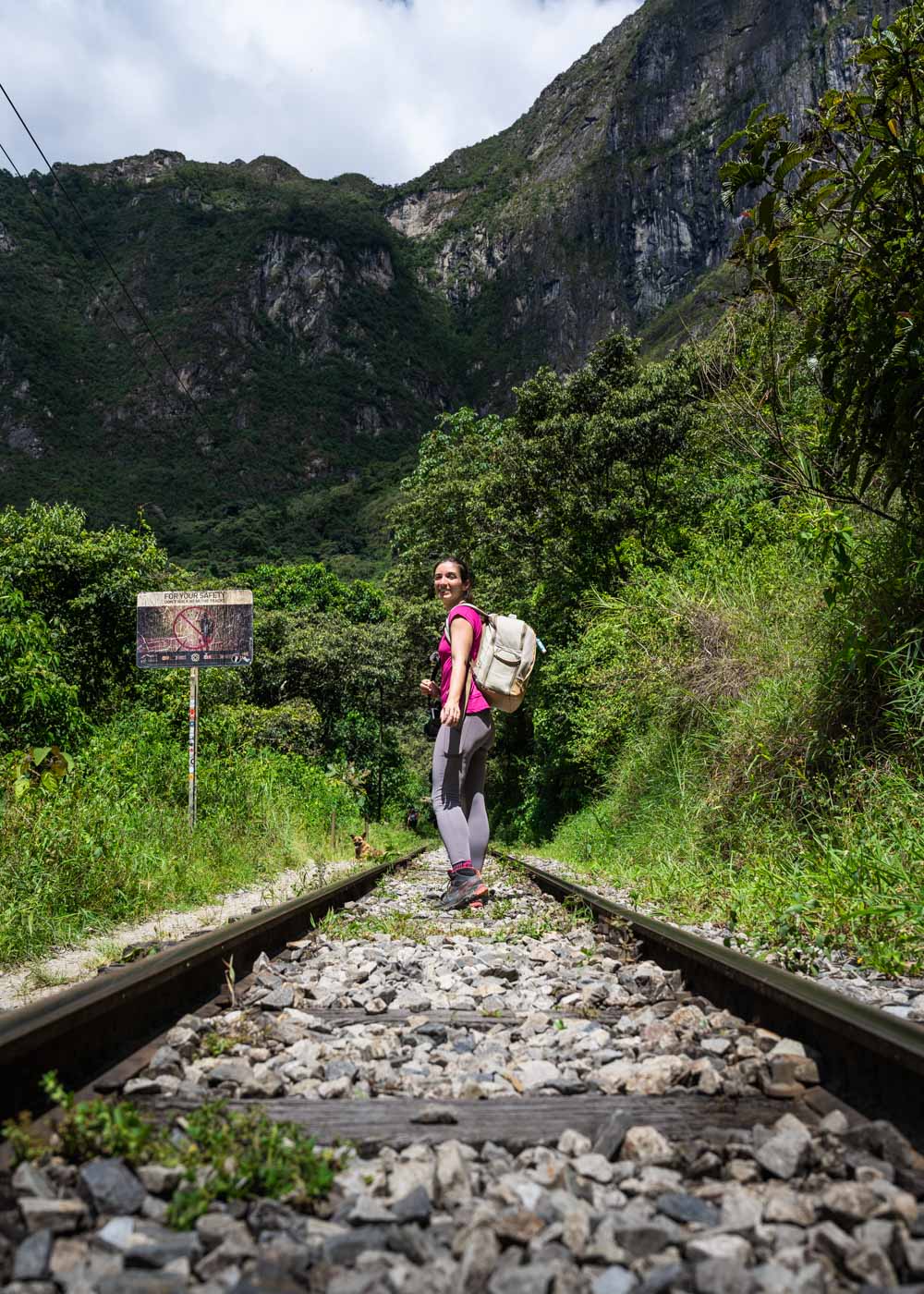 Sara in hiking gear and a backpack posing on a train track surrounded by trees, mountains and a run-down sign.
