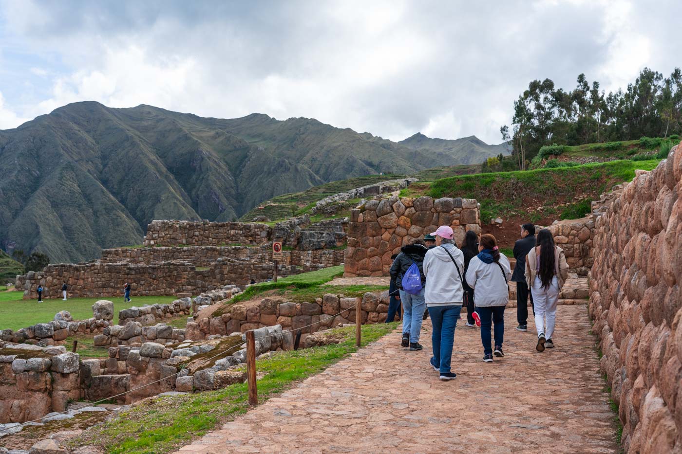 A group of tourists walking down a path inbetween the walls of Chinchero archeological site with a view of mountains in the Sacred valley of Peru.