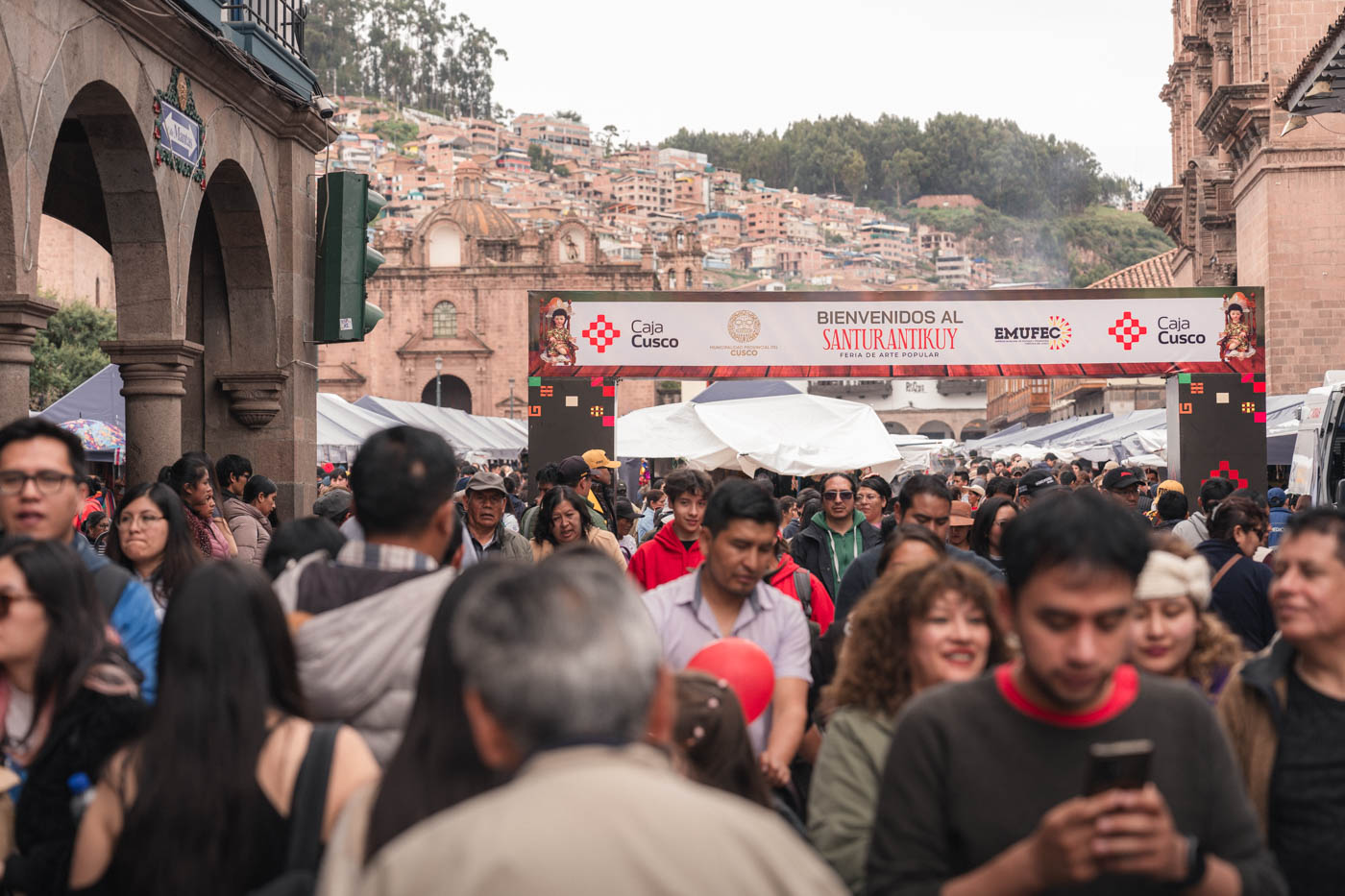 A crowd of people entering and exiting under a banner for the Santurantikuy Arts Festival in Plaza de Armas in Cusco.
