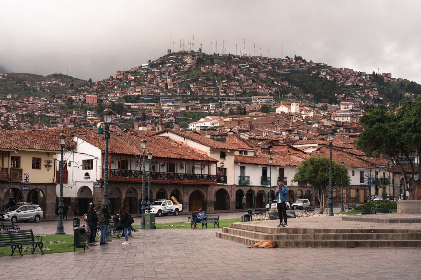 People and street dogs going about their morning on an overcast day in Plaza de Armas with a view of Cusco built up the side of a mountain.