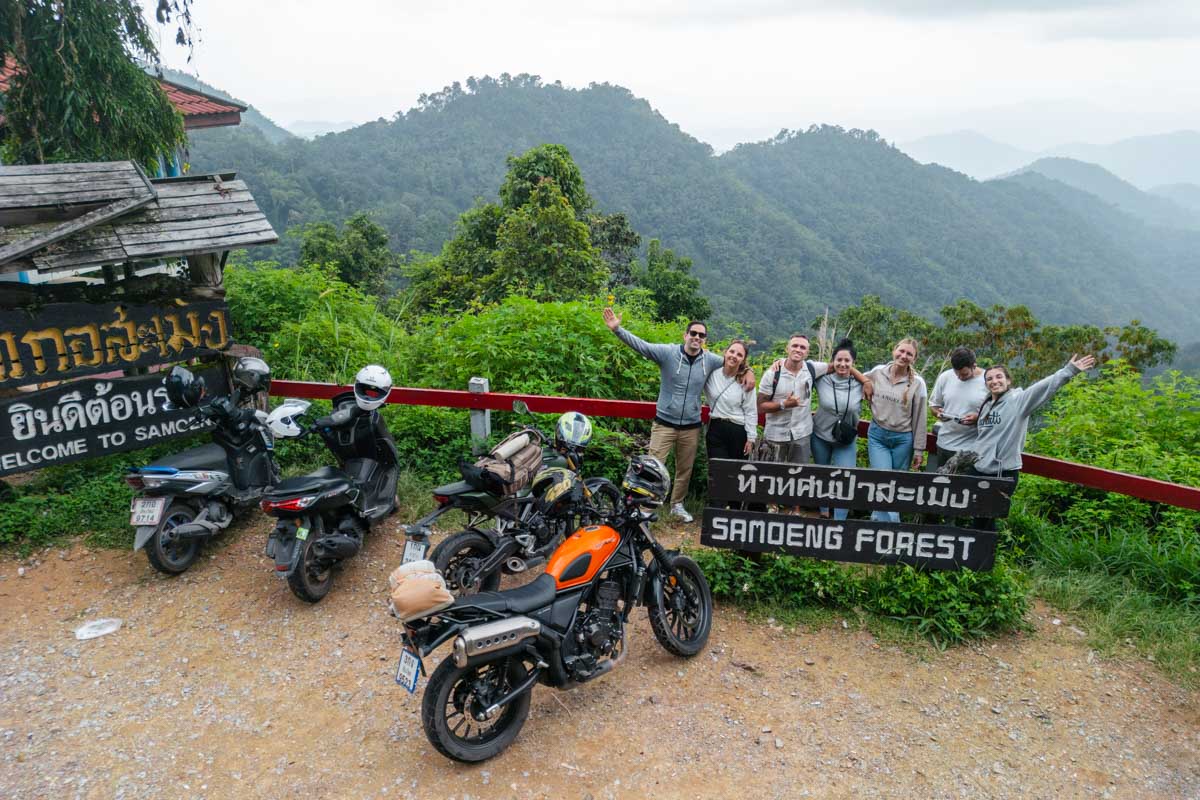 A group photo of us posing with our friends beside the Samoeng Forest sign, with a stunning view of mountains and forests in distance and our motorbikes parked to our left.