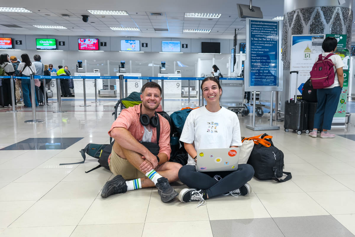 Ryan and Sara sitting in the middle of Chiang Mai International airport posing with a laptop while people behind check into their flight.