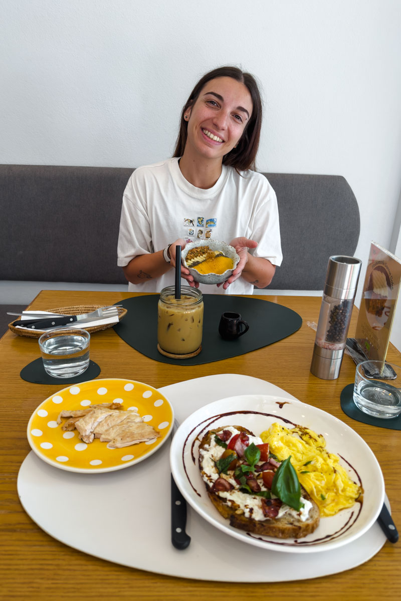 Sara, inside Healthy Junk, looking very happy while holding a bowl of yogurt and fruit, beside a table with burrata toast, eggs, and chicken on it.