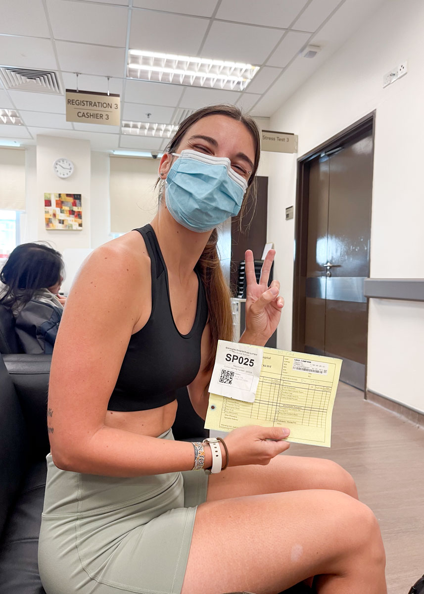 Sara throwing up a peace sign while holding a hospital document and wearing a blue surgical mask in Gleneagles Hospital in Kuala Lumpur.