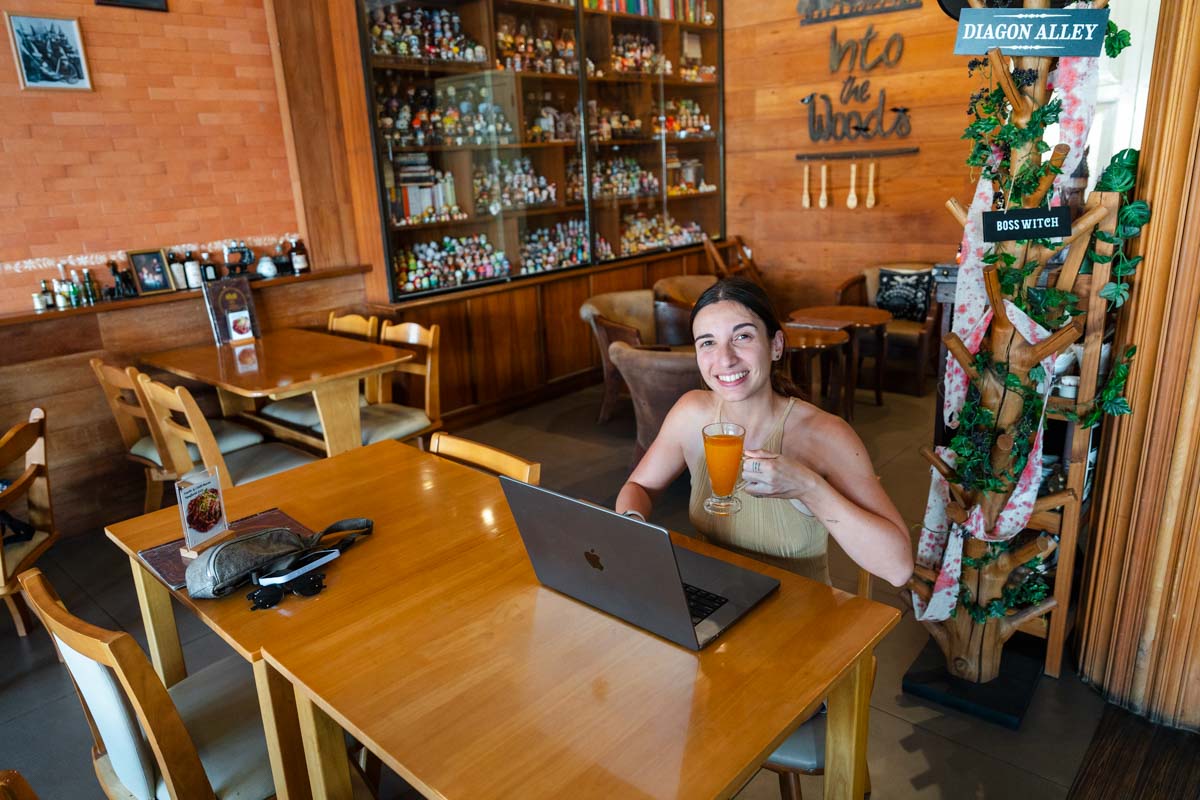 Sara sitting at a table in Into the Woods cafe, holding an orange drink while working at a laptop. Around her are witch-themed signs and small figurines in a glass case.