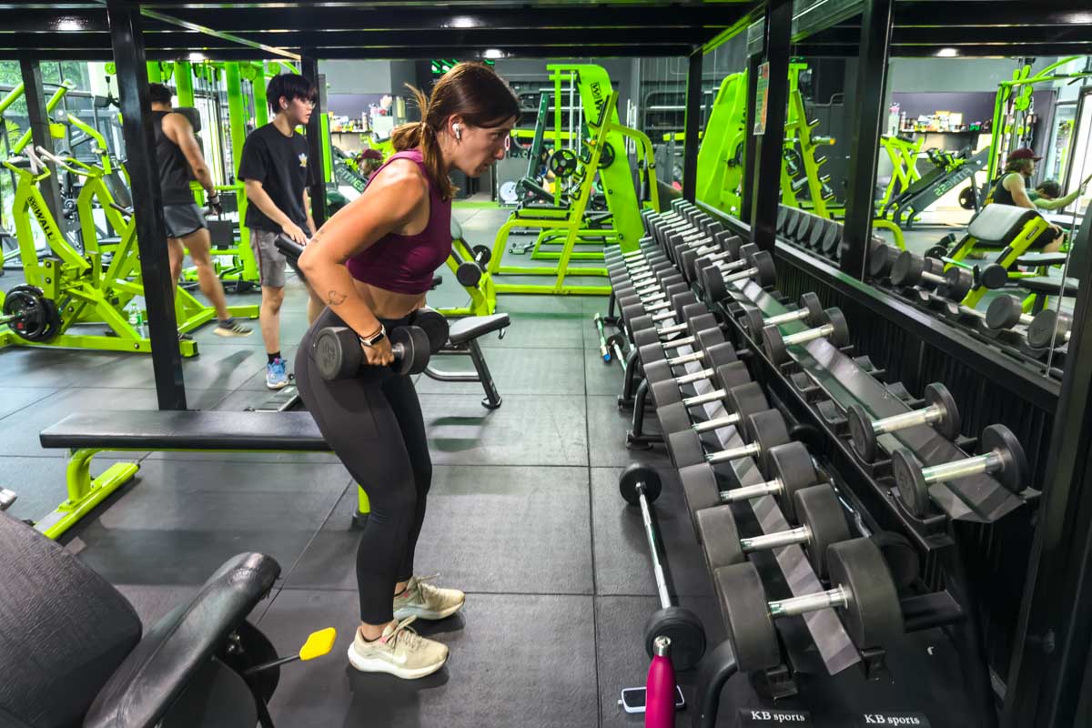 Sara standing in the free weights sections of The Wall gym, lifting two dumbbells with a concentrated look on her face. Behind her are people working out on green gym machines.
