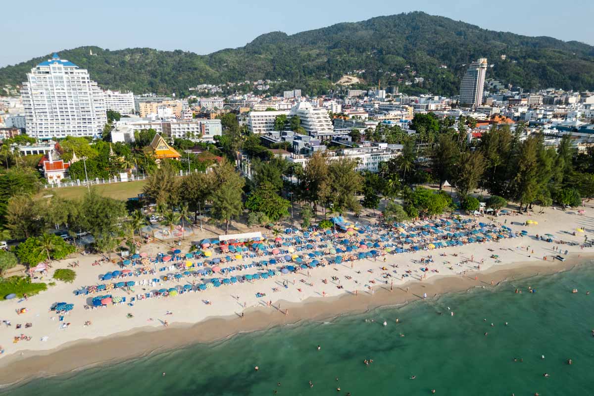 An aerial view over Patong Beach in Phuket. It's super busy, there are lots of people swimming, rows of umbrellas on the beach, and lots of hotels behind.
